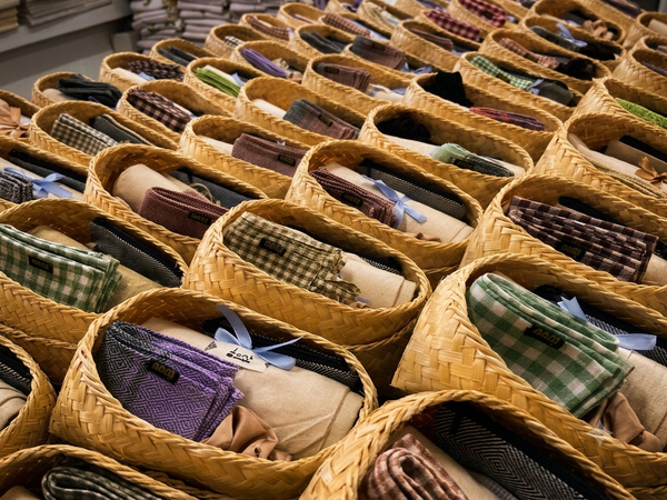 Rows of woven straw baskets filled with folded clothing and accessories, including plaid shirts, purple fabric items, and rolled garments, arranged in a market-style display.