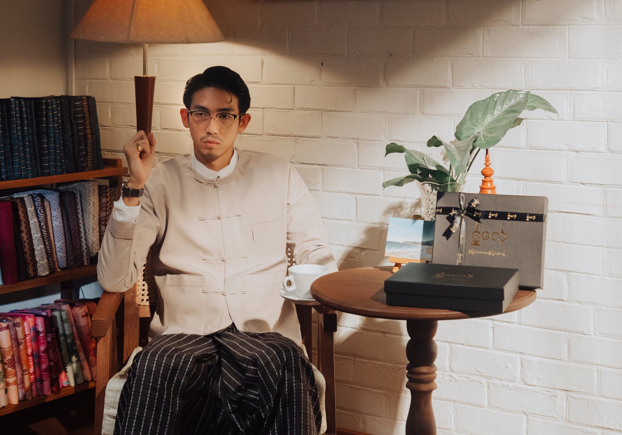 Man wearing Dalae Tite Pon ( traditional jacket ) sitting in a chair holding a glass, with a table and books in the background